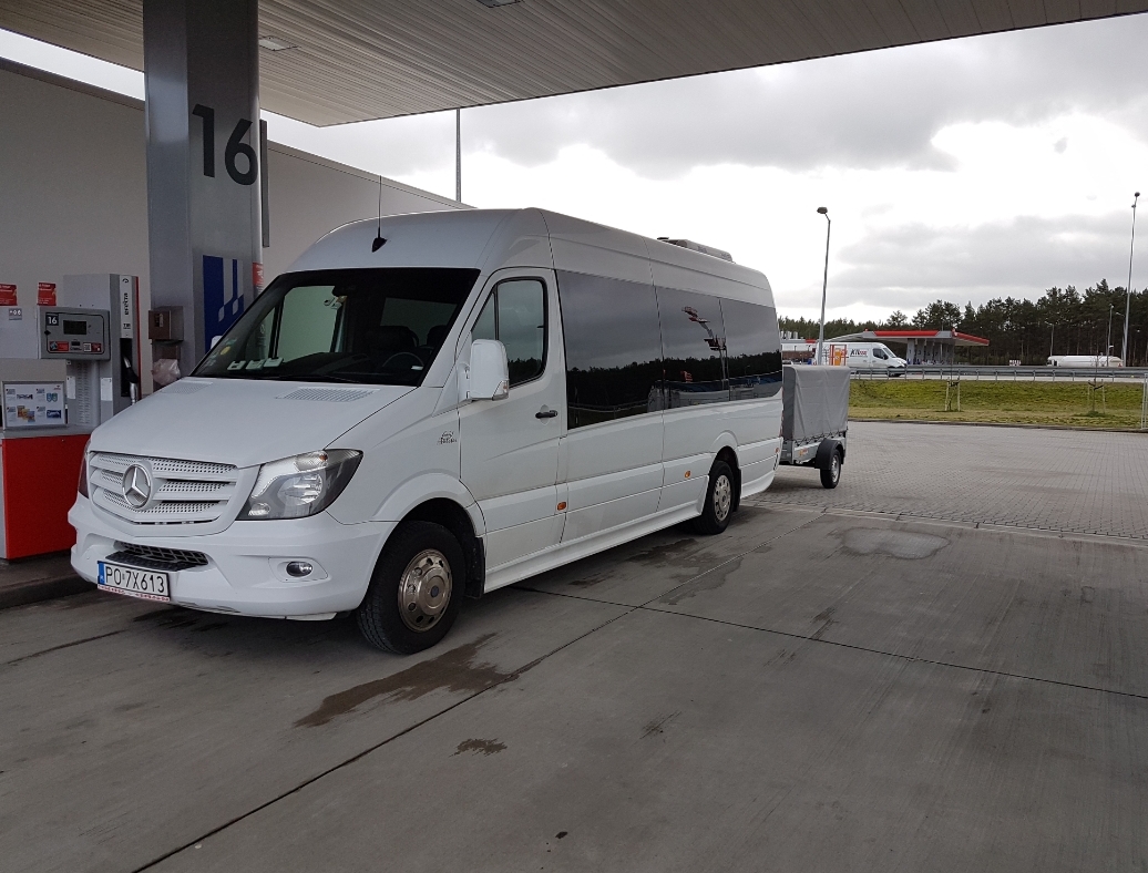 Biały Mercedes Sprinter z przyczepką na stacji benzynowej, widok z przodu i boku, pochmurne niebo w tle.