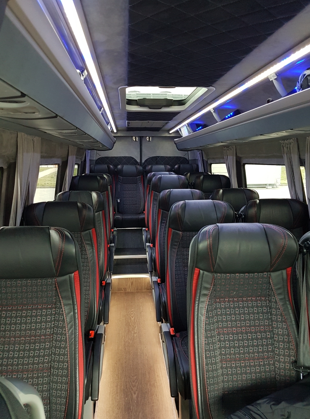 Wnętrze luksusowego busa z czarnymi skórzanymi fotelami z czerwonymi akcentami, oświetleniem LED i jasną podłogą.
