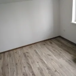 Pomalowany na biało pokój z ułożonymi panelami podłogowymi, puszka z farbą i pędzlem przy grzejniku.