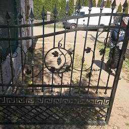 Usługi ślusarsko spawalnicze - Otwarta, czarna, metalowa brama z motywem winogron i greckim wzorem, zamontowana przy ogrodzeniu z siatki, w tle biały samochód dostawczy.