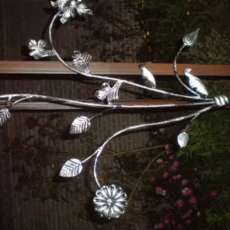 Dekoracyjny element metalowy z motywem kwiatowym i ptaków, przymocowany do ramy, z widocznym tłem roślinnym.