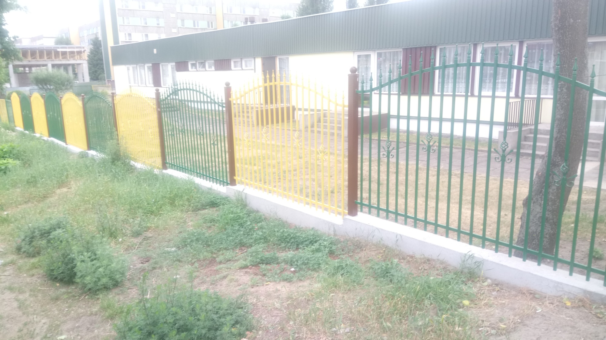Metalowe ogrodzenie z elementami dekoracyjnymi, pomalowane na zielono i żółto, otaczające budynek z zielonym dachem.