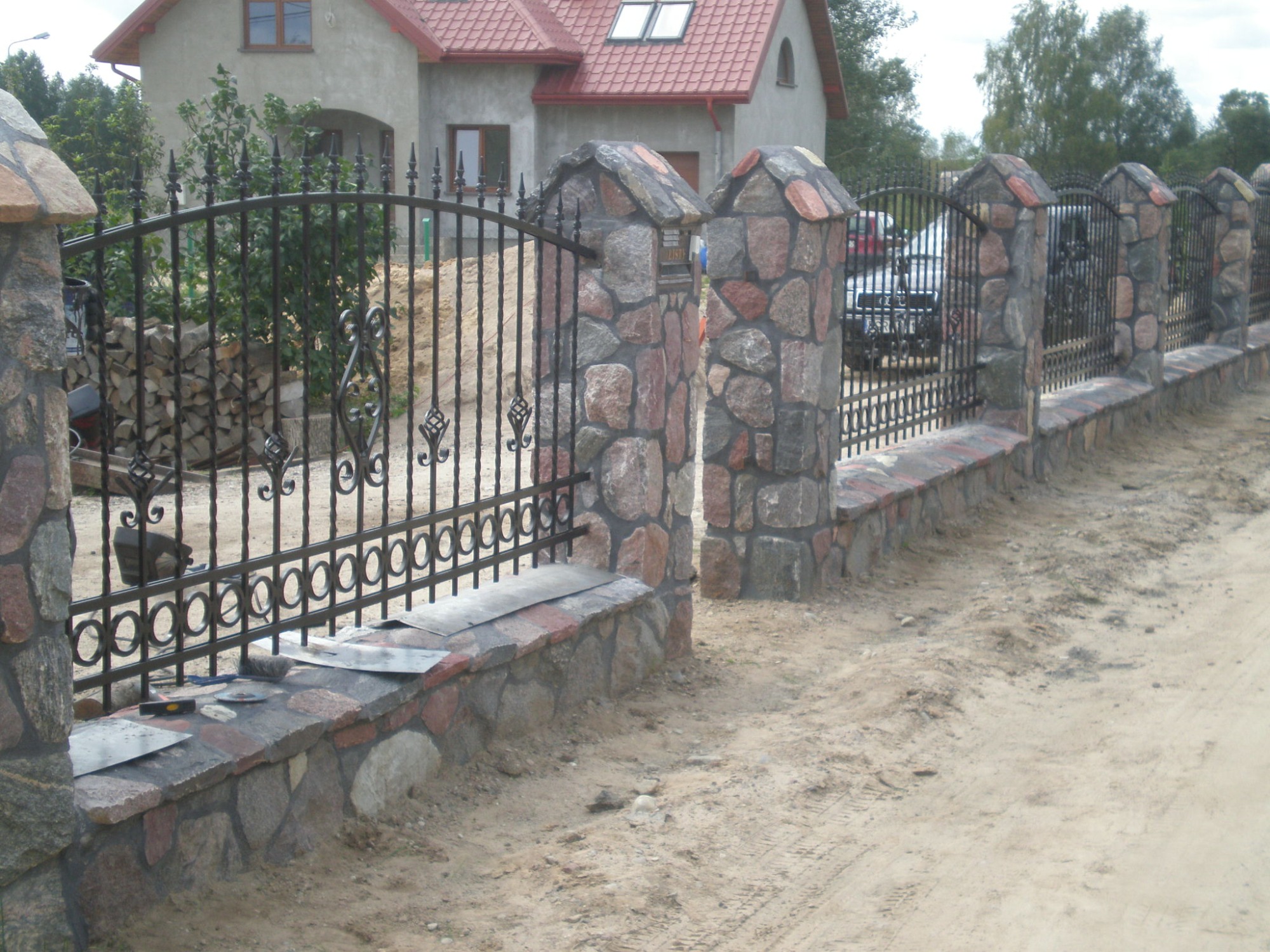 Ozdobna, czarna, metalowa brama z kamiennymi słupkami i podmurówką, w tle dom z czerwoną dachówką i stos drewna