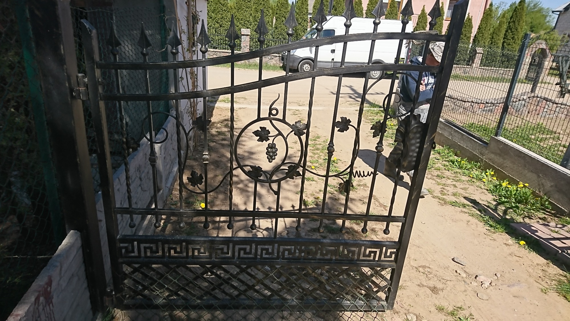 Otwarta, czarna, metalowa brama z motywem winogron i greckim wzorem, zamontowana przy ogrodzeniu z siatki, w tle biały samochód dostawczy.