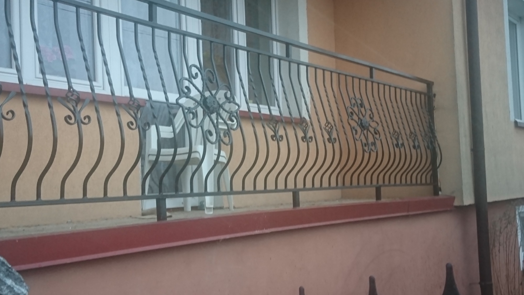 Metalowa balustrada balkonowa z dekoracyjnymi elementami kwiatowymi, widoczna z zewnątrz budynku.