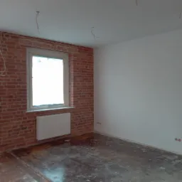 Wnętrze pokoju w trakcie remontu: ściana z surowej czerwonej cegły z oknem i grzejnikiem, biała pomalowana ściana, odsłonięta podłoga z desek, wystające kable z sufitu.