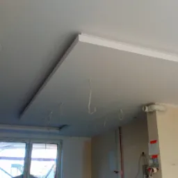 Niedokończony sufit podwieszany z wystającymi przewodami elektrycznymi w jasnym pomieszczeniu z oknem i instalacją wentylacyjną.