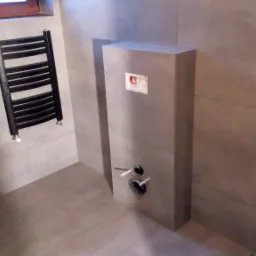 Nowoczesna łazienka z szarymi płytkami, zabudową stelaża WC, grzejnikiem drabinkowym w kolorze czarnym i fragmentem drewnianej okładziny ściany.