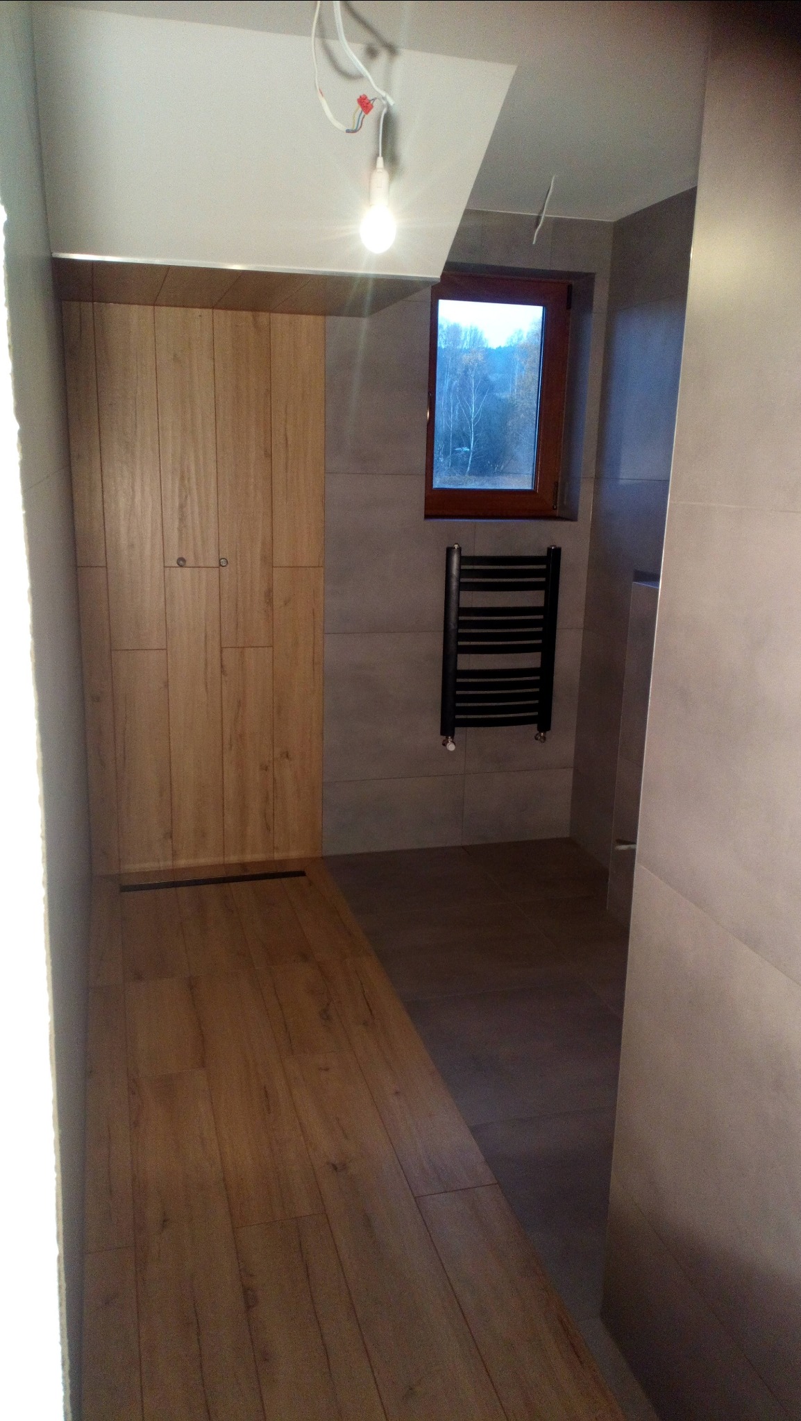 Nowoczesna łazienka z drewnopodobną podłogą i kabiną prysznicową z drzwiami z paneli drewnianych, grzejnikiem drabinkowym i oknem z widokiem na jesienne drzewa.