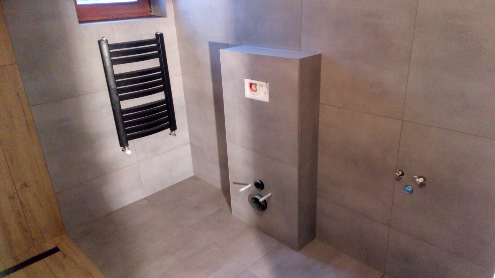 Nowoczesna łazienka z szarymi płytkami, zabudową stelaża WC, grzejnikiem drabinkowym w kolorze czarnym i fragmentem drewnianej okładziny ściany.