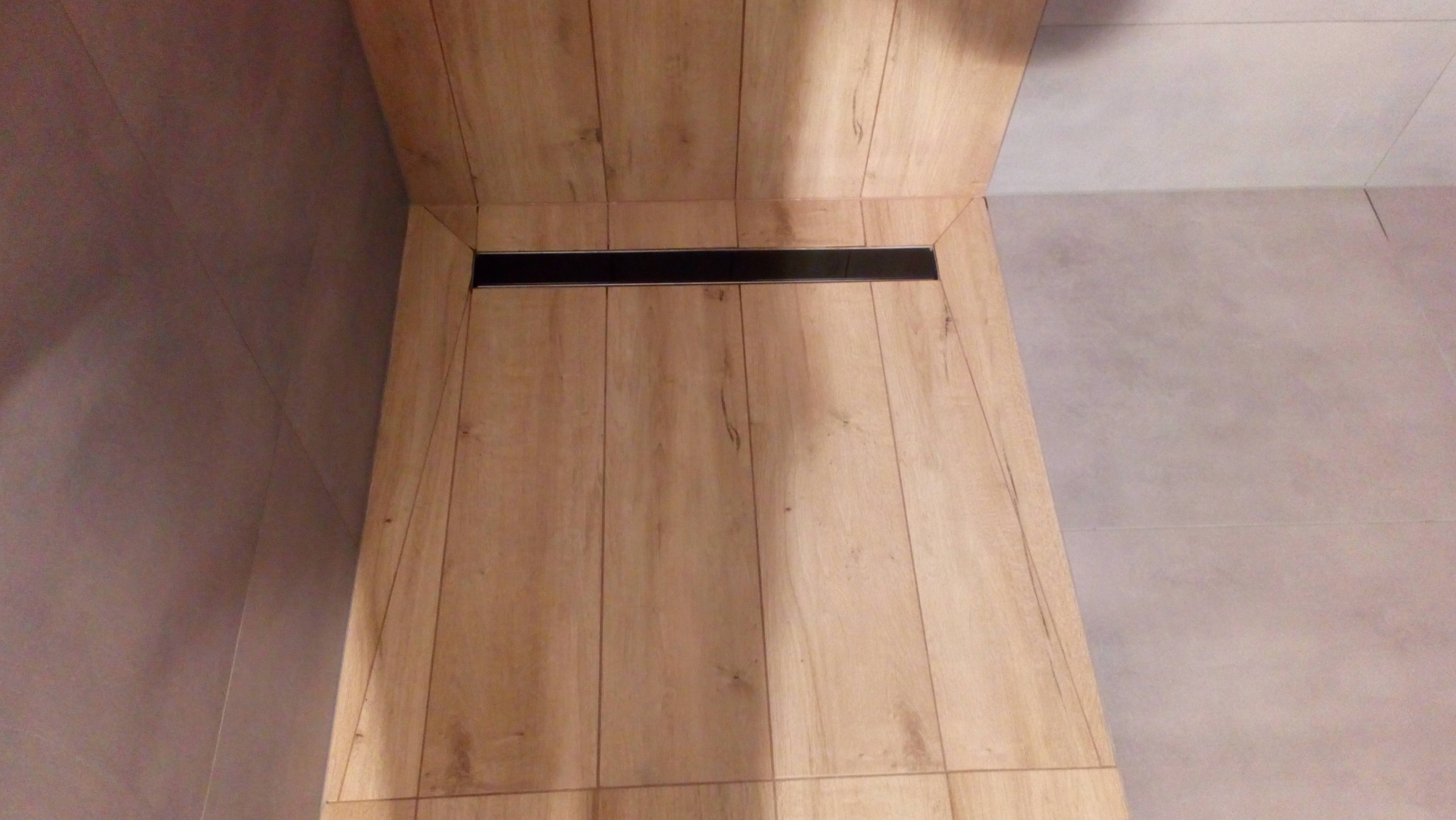 Wykończony brodzik prysznicowy z odpływem liniowym, wyłożony płytkami imitującymi drewno, sąsiadujący z szarą płytką ścienną.