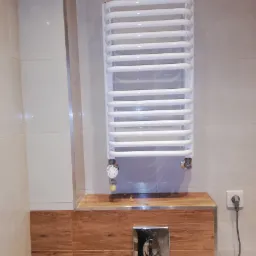 Biały, drabinkowy grzejnik łazienkowy zamontowany na ścianie z beżowych płytek, obok drewnopodobnej zabudowy ze srebrnym przyciskiem spłuczki i gniazdkiem elektrycznym.