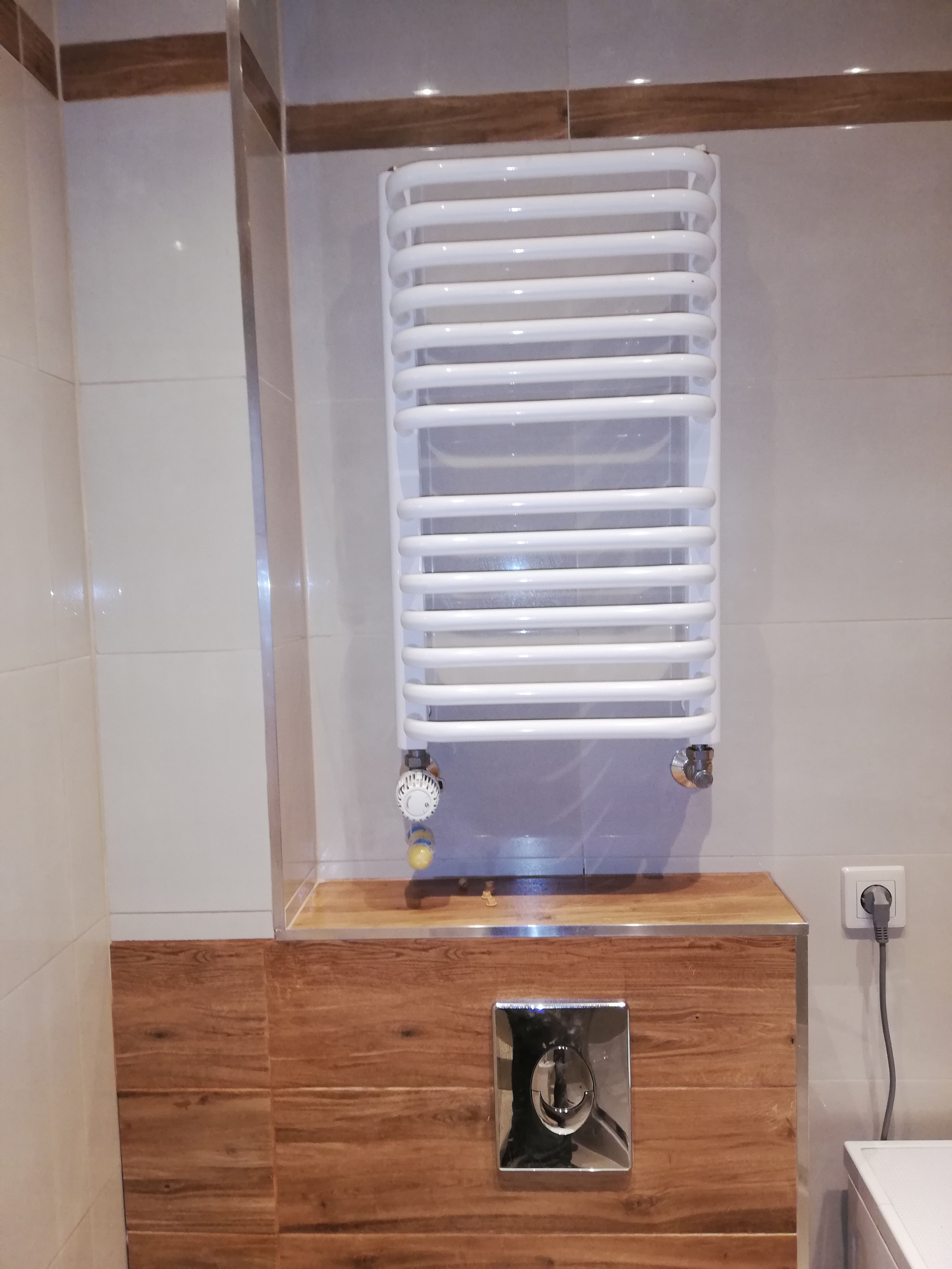 Biały, drabinkowy grzejnik łazienkowy zamontowany na ścianie z beżowych płytek, obok drewnopodobnej zabudowy ze srebrnym przyciskiem spłuczki i gniazdkiem elektrycznym.