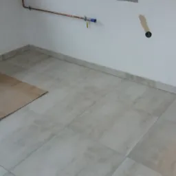 Jasne, kwadratowe płytki podłogowe ułożone w pomieszczeniu, z widocznymi instalacjami hydraulicznymi na ścianie i kartonem ochronnym na podłodze.