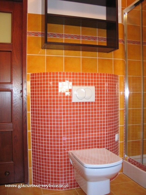 Wykończona łazienka z pomarańczowymi płytkami i mozaiką wokół toalety, wisząca szafka na ścianie i częściowo widoczny prysznic za szklaną ścianką.