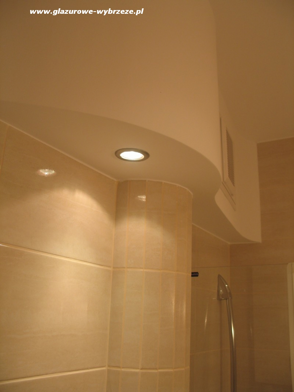 Wykończenie łazienki z beżowymi płytkami, okrągłym oświetleniem w suficie podwieszanym i widoczną kabiną prysznicową.