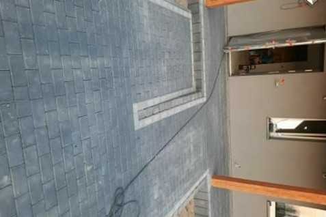 Ułożona szara kostka brukowa z jasnymi obrzeżami i widocznym kablem na powierzchni, fragment budynku w tle.