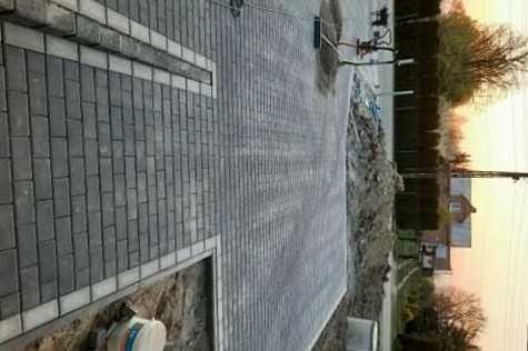 Świeżo ułożona, szara kostka brukowa na podjeździe, z widocznym fragmentem chodnika obramowanego jaśniejszą kostką, na dalszym planie maszyna do ubijania i pryzma ziemi.