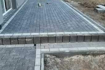 Fragment nowo ułożonej nawierzchni z kostki brukowej w odcieniach szarości, z widocznym obrzeżem i fragmentem przygotowanego podłoża z piasku.