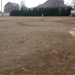 Przygotowany teren pod trawnik z widocznymi śladami po niwelacji i wytyczonymi okręgami, w tle domy i drzewa.