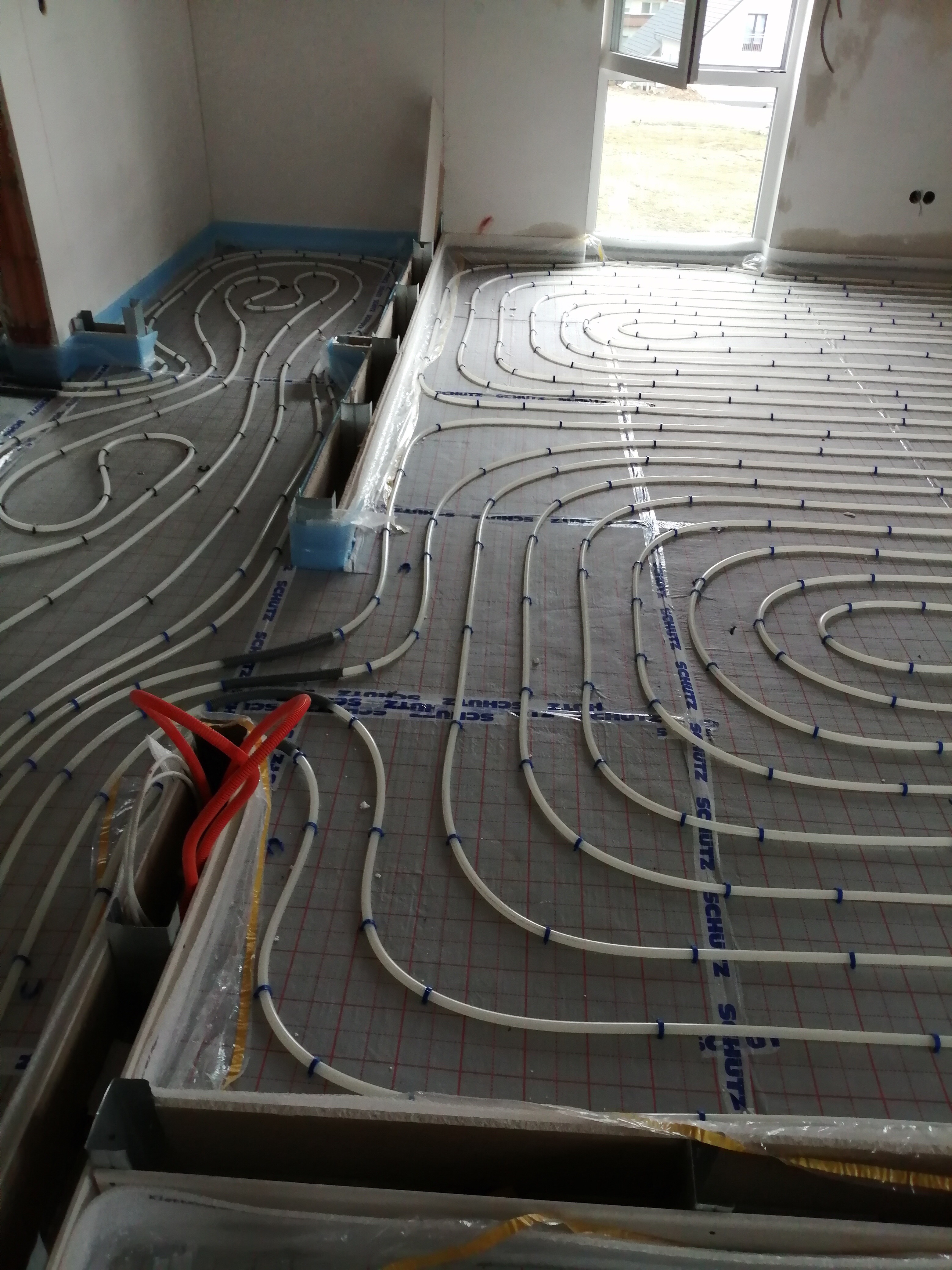 Rozłożone białe rury ogrzewania podłogowego na siatce z folią aluminiową, widoczne nieotynkowane ściany i okno, przygotowanie do wylewki.