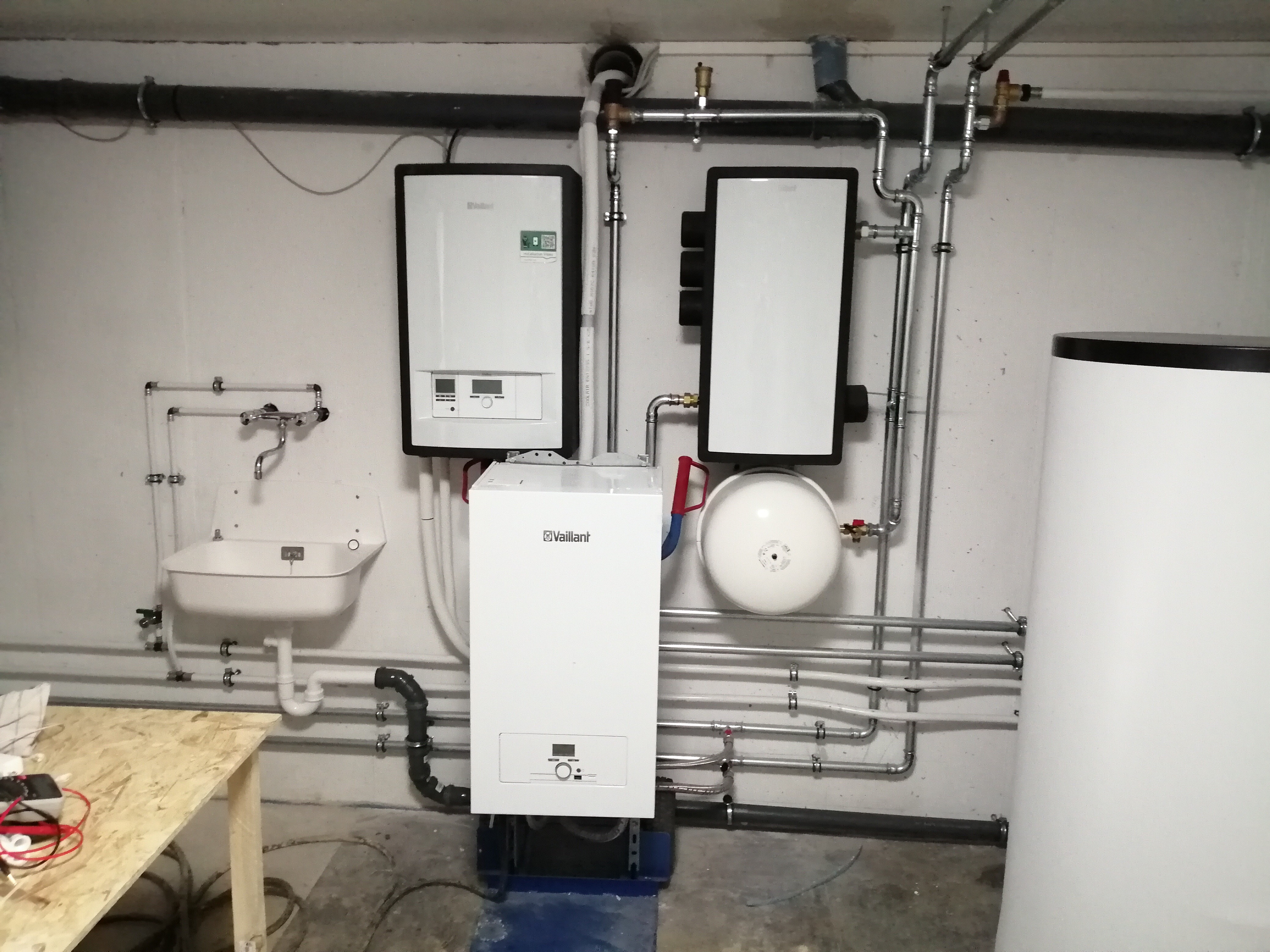 Kompaktowa kotłownia w piwnicy z trzema kotłami grzewczymi Vaillant, zbiornikiem buforowym i instalacją hydrauliczną z rur miedzianych, z widocznym zlewem warsztatowym i prowizorycznym stołem.