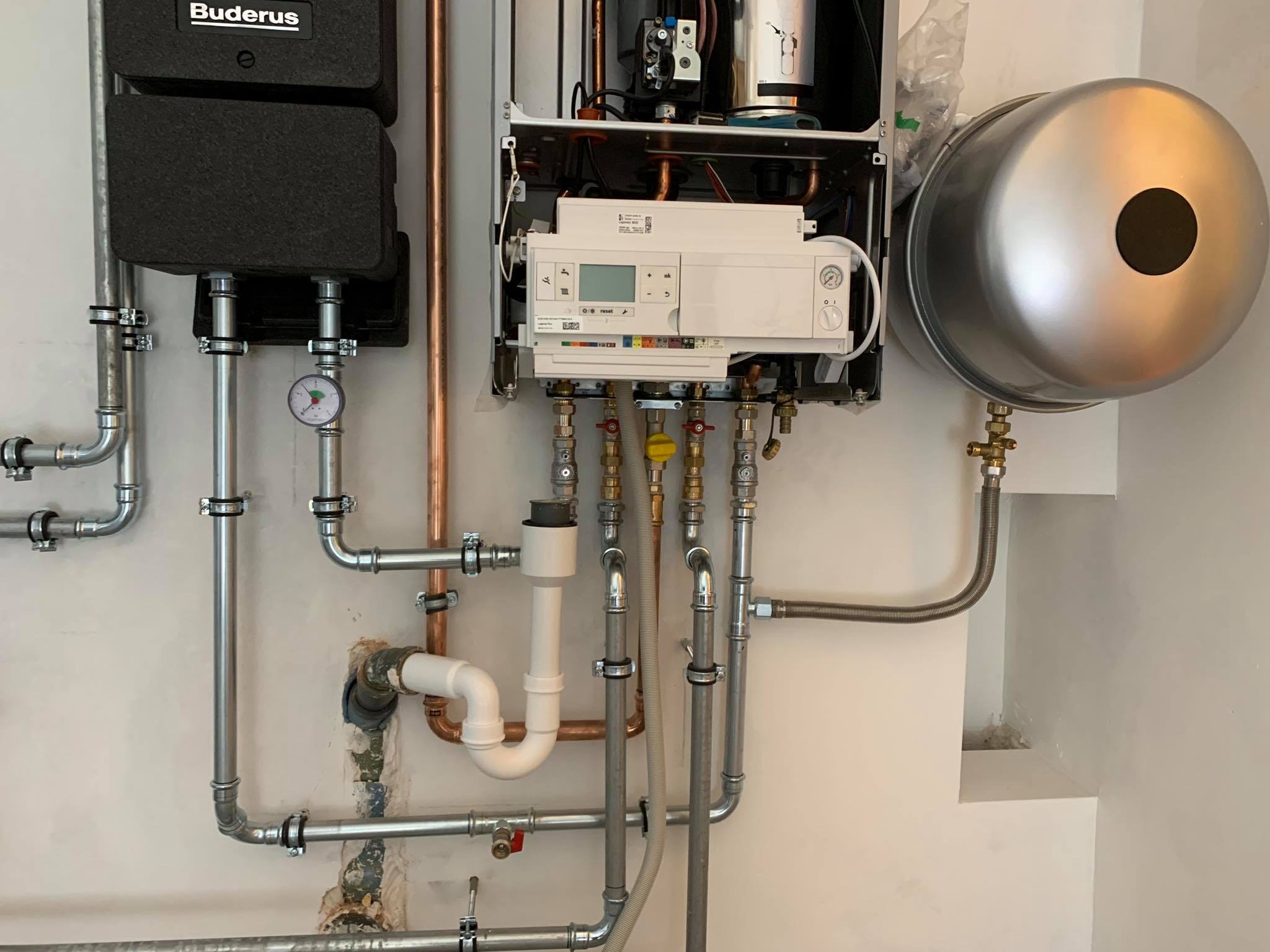 Złożona instalacja kotła grzewczego Buderus z zasobnikiem wody, widoczne liczne rury miedziane i stalowe, manometr i sterownik elektroniczny, montaż na ścianie o nierównej powierzchni.
