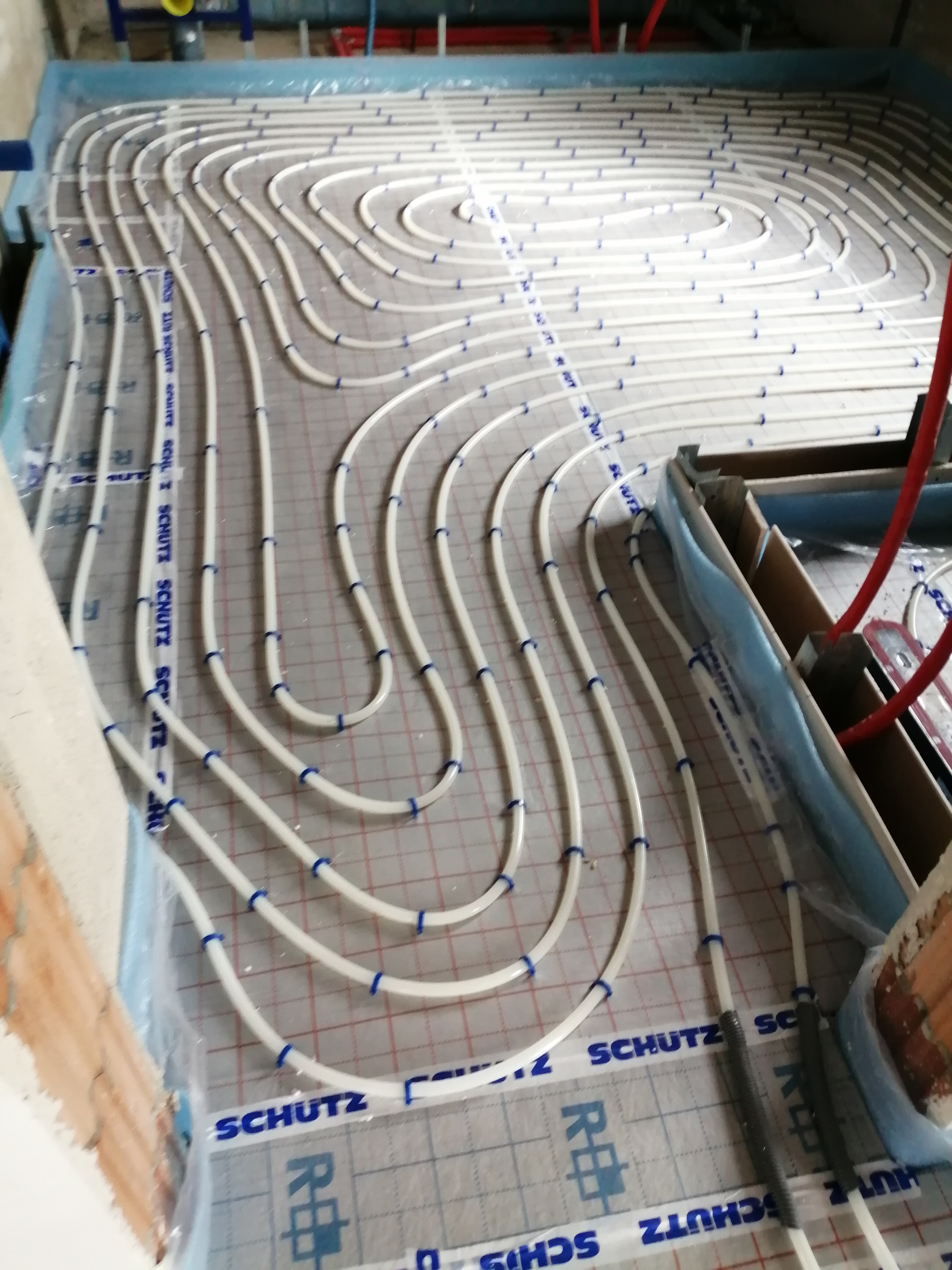 Białe rury ogrzewania podłogowego, mocowane niebieskimi uchwytami do folii z nadrukiem SCHUTZ, ułożone spiralnie na przygotowanej powierzchni w trakcie montażu.