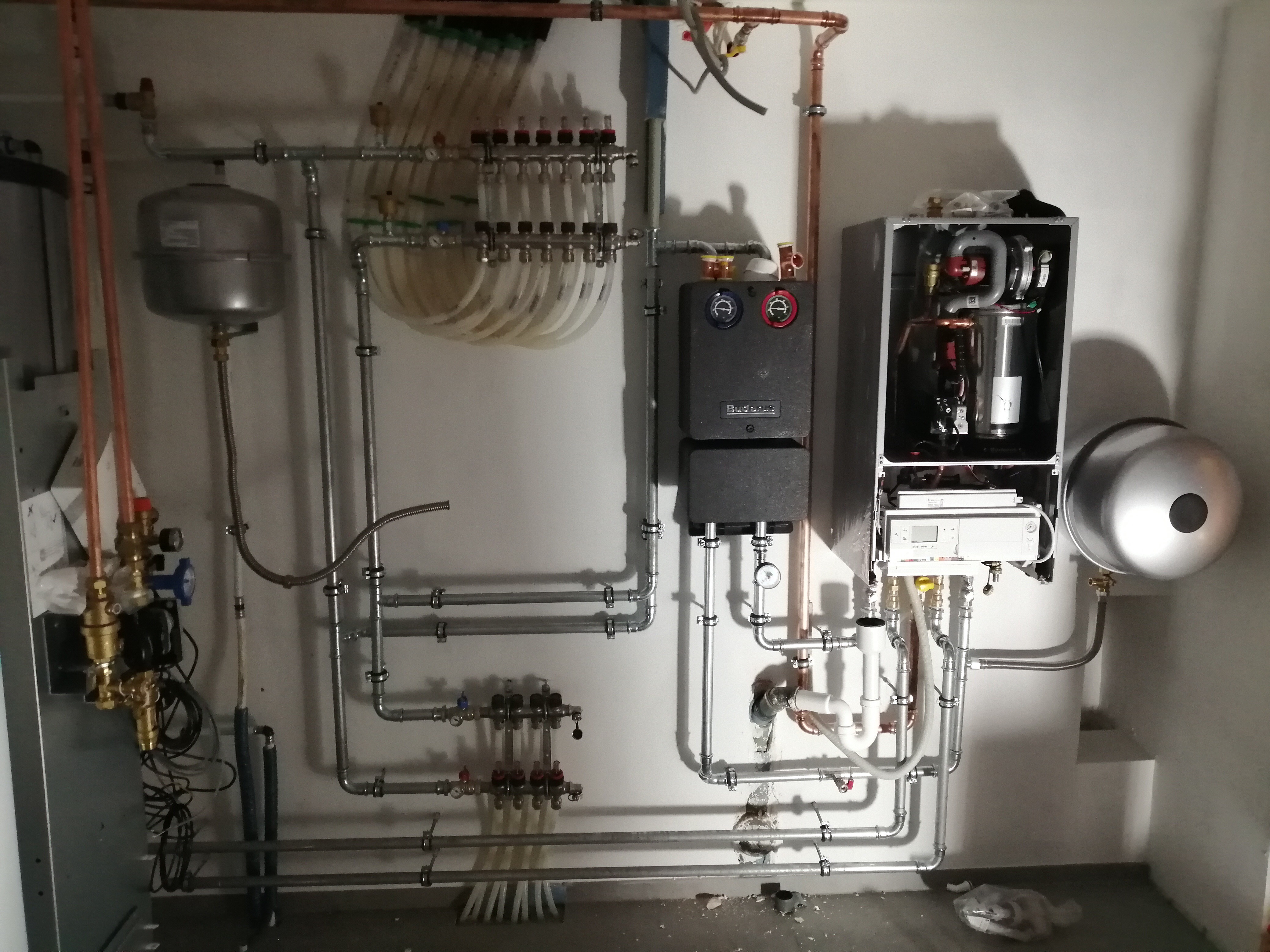 Rozbudowana instalacja centralnego ogrzewania z kotłem gazowym, rozdzielaczami i naczyniem wzbiorczym, widoczne rury miedziane i stalowe, manometry i zawory, w surowym stanie wykończenia.