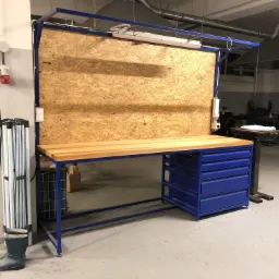 Stół warsztatowy z drewnianym blatem, niebieską ramą i szafką z szufladami, na tle ściany z płyty OSB, oświetlony lampą fluorescencyjną, z gumowymi butami i rozkładanym stojakiem obok.