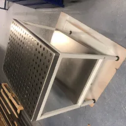 Stół spawalniczy z perforowanym blatem i białym metalowym stelażem, widoczny w warsztatowym otoczeniu z paletą i metalowymi profilami w tle.