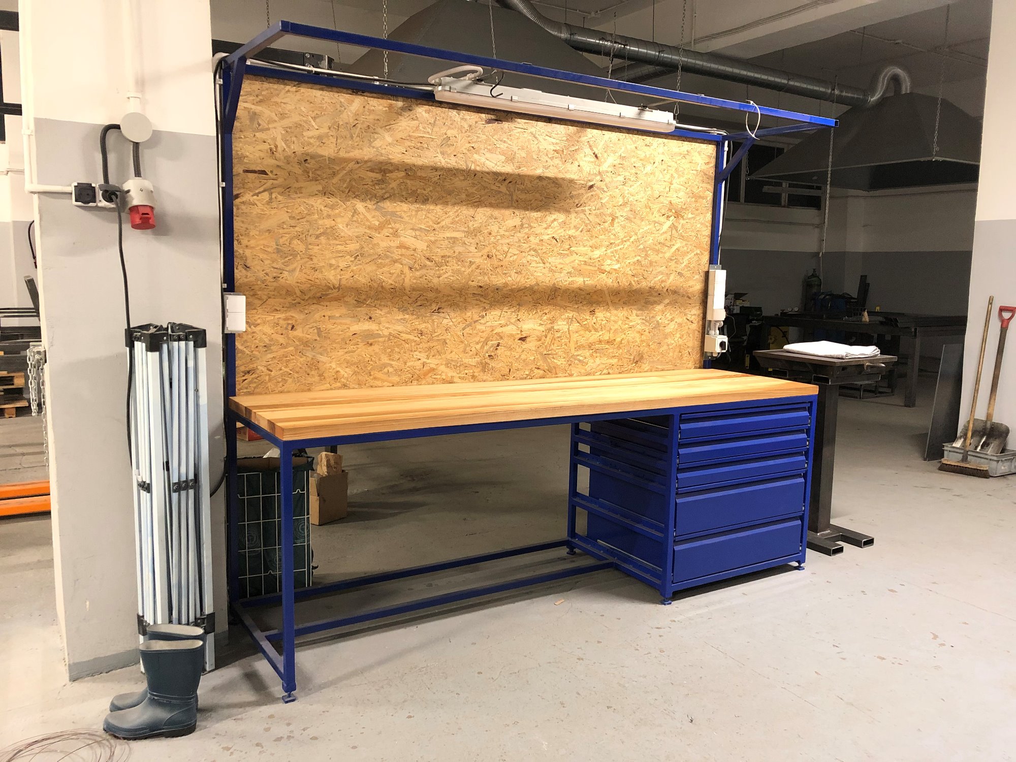 Stół warsztatowy z drewnianym blatem, niebieską ramą i szafką z szufladami, na tle ściany z płyty OSB, oświetlony lampą fluorescencyjną, z gumowymi butami i rozkładanym stojakiem obok.