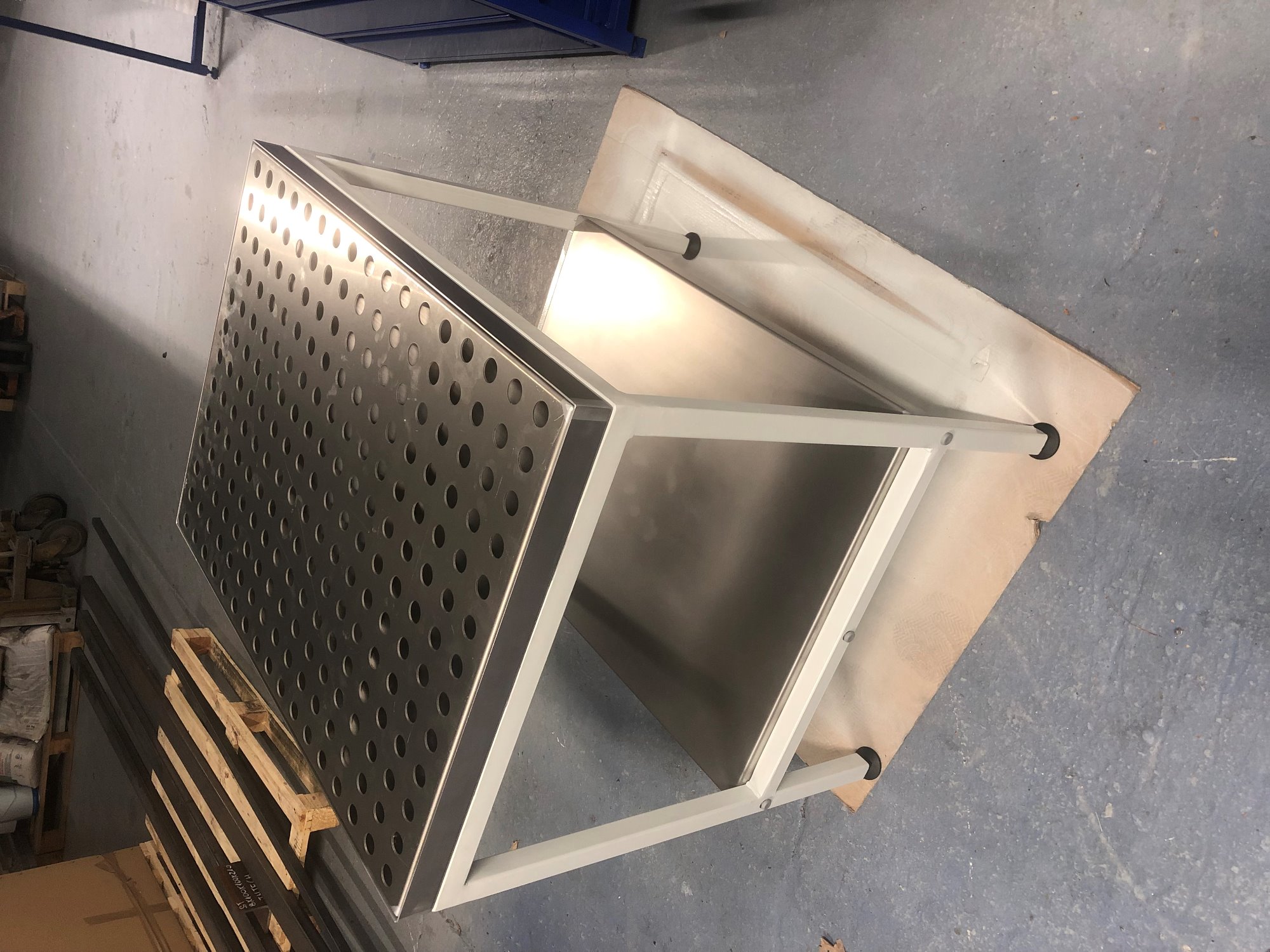 Stół spawalniczy z perforowanym blatem i białym metalowym stelażem, widoczny w warsztatowym otoczeniu z paletą i metalowymi profilami w tle.