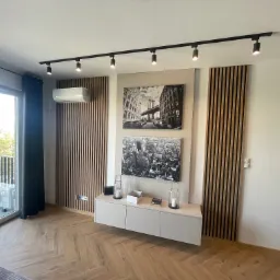 Nowoczesny salon z klimatyzacją na ścianie z drewnianymi lamelami, czarne reflektory sufitowe, obrazy Nowego Jorku i jednostka zewnętrzna klimatyzatora na balkonie.