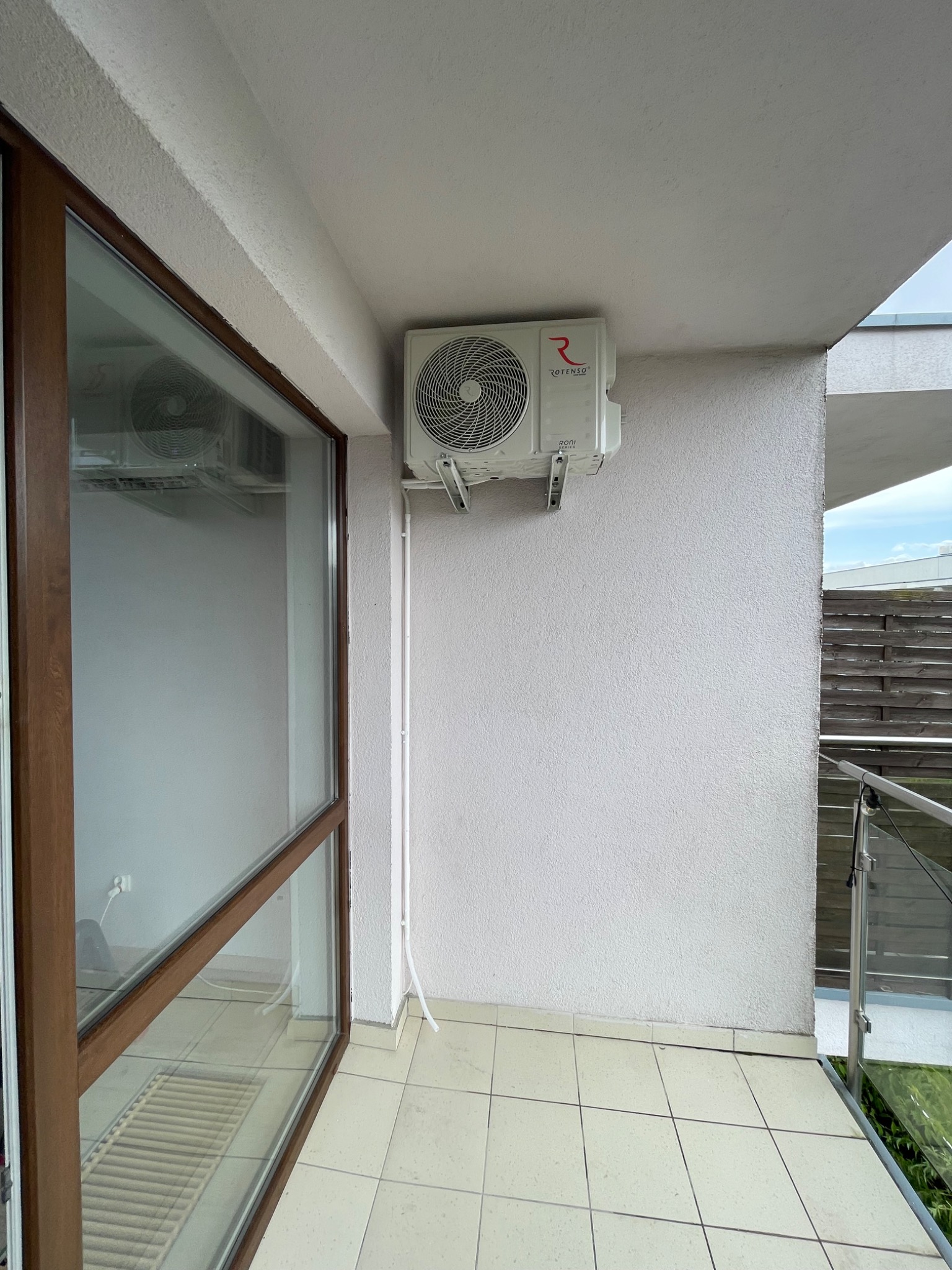 Klimatyzator Rotenso zamontowany na ścianie balkonu, widoczne białe rurki instalacyjne i fragment okna z brązową ramą. Widok na zewnątrz z drewnianym ogrodzeniem w tle.
