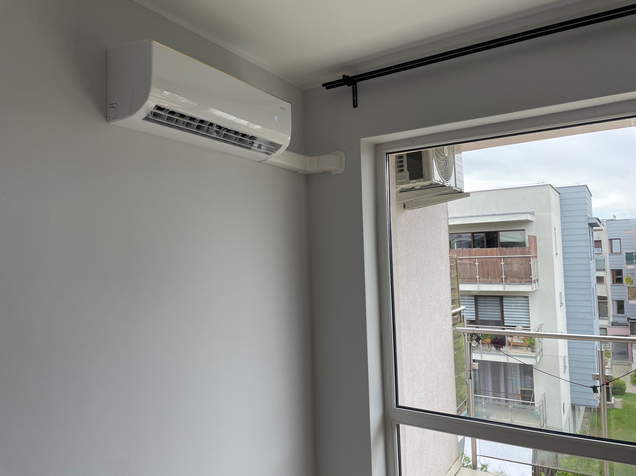 Biała klimatyzacja ścienna zamontowana w jasnym pokoju, widok na balkon zza okna. Widoczne rury odprowadzające. Minimalistyczny design.