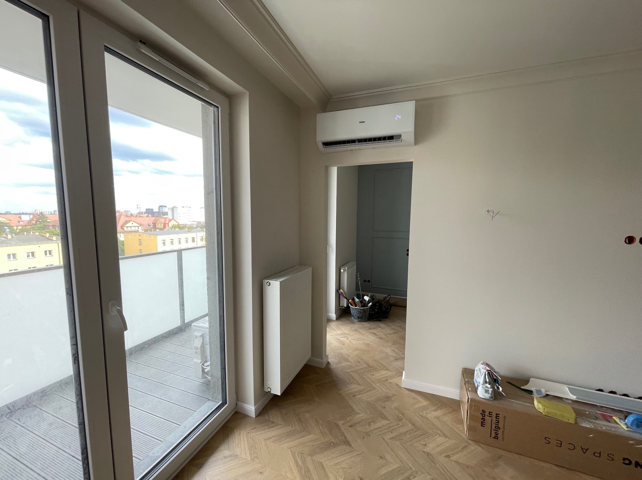 Biały klimatyzator Haier zamontowany na ścianie w jasnym pokoju z parkietem w jodełkę, balkon z widokiem na miasto i grzejnik.