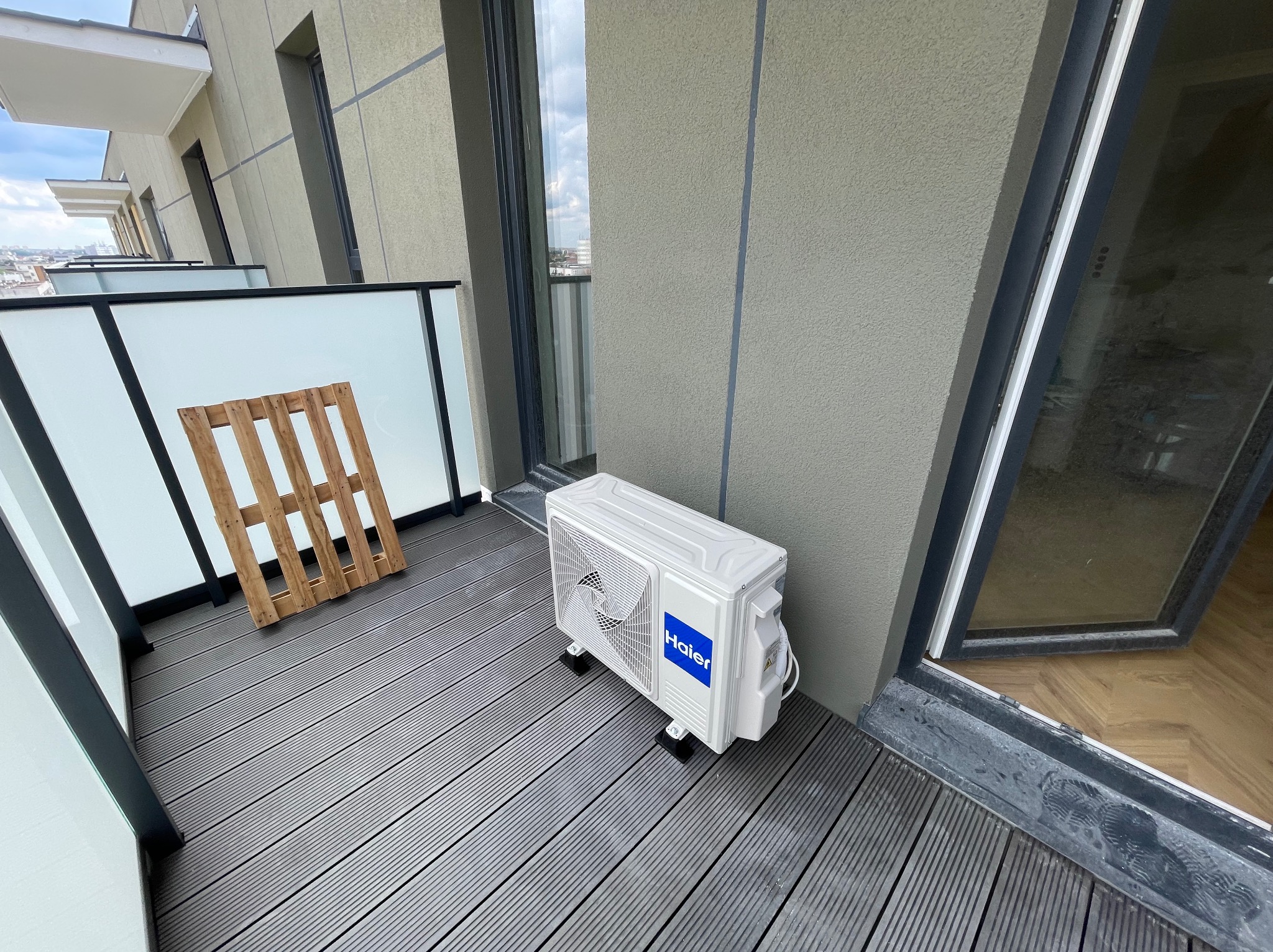 Biały klimatyzator Haier na szarym tarasie z deską kompozytową. Widoczny fragment balkonu z paletą. Montaż klimatyzacji na balkonie w nowoczesnym budownictwie.