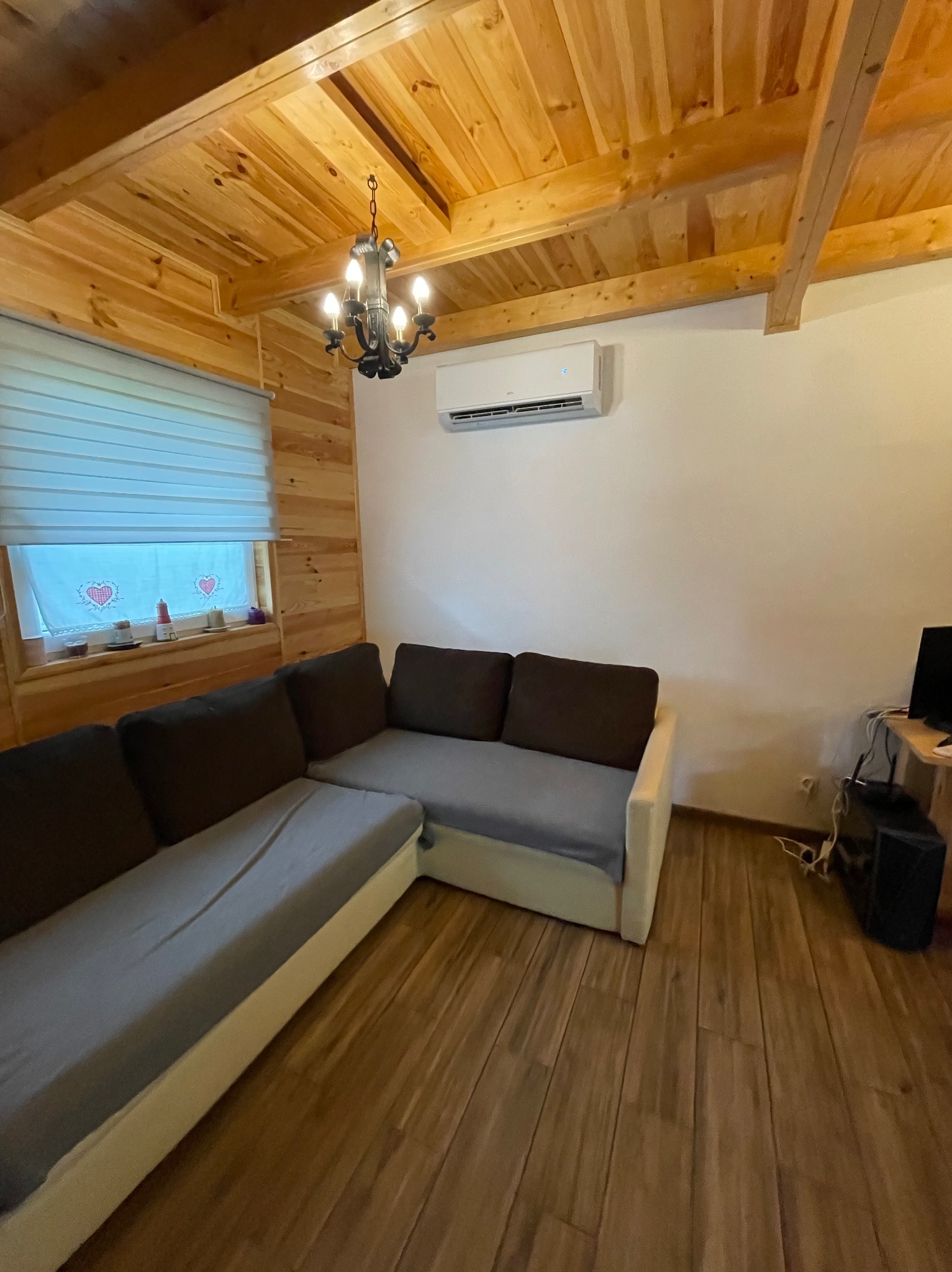 Klimatyzator ścienny w drewnianym salonie z oknem, żaluzjami i kanapą narożną. Widoczne drewniane panele na ścianach i suficie, oraz ozdobny żyrandol.