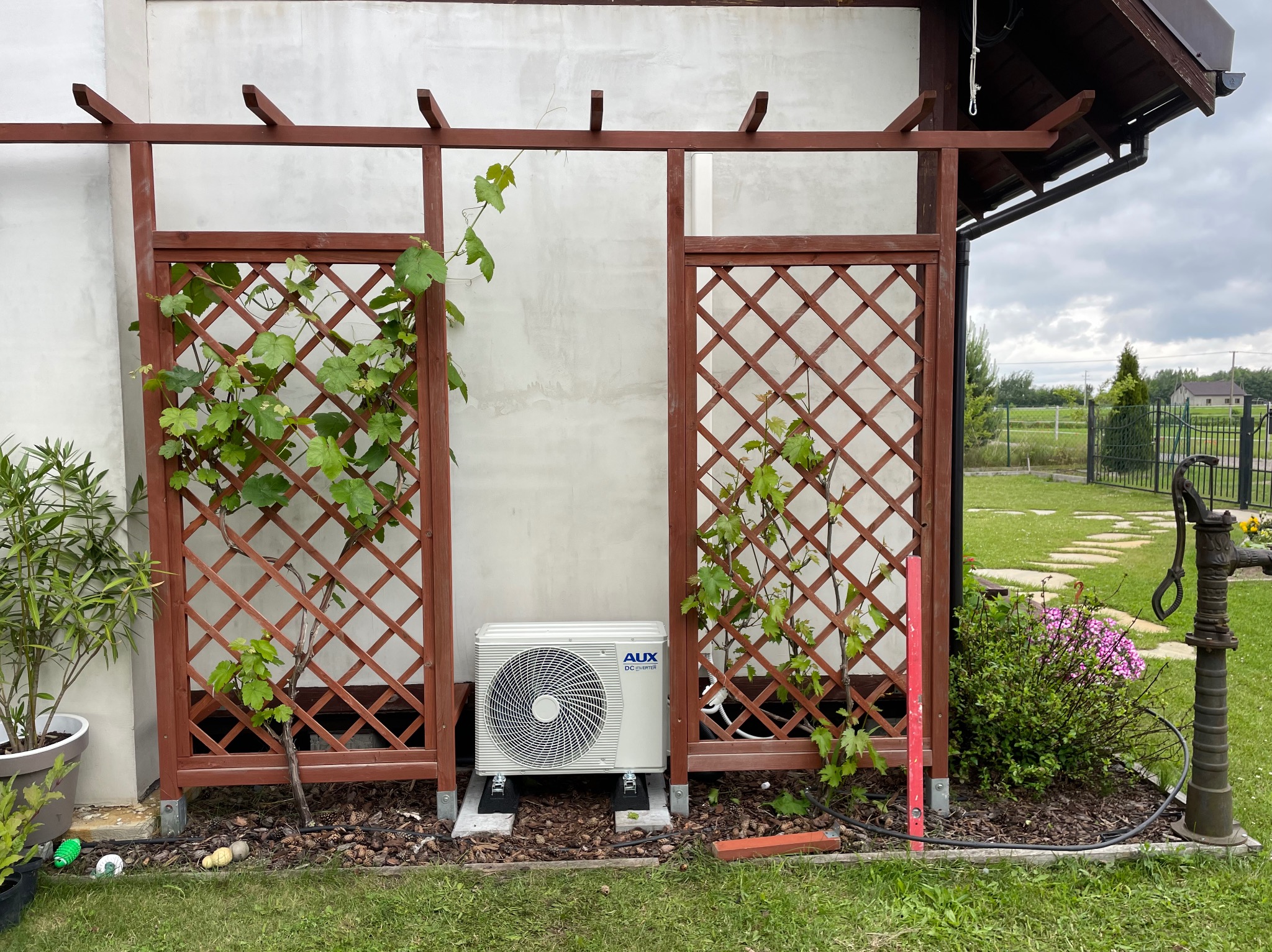 Biała jednostka zewnętrzna klimatyzacji AUX na betonowych podstawach, umieszczona pod drewnianą pergolą z pnącymi roślinami w ogrodzie.