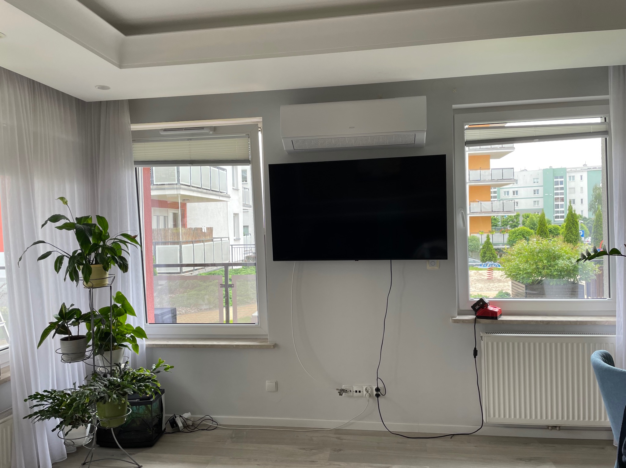 Biała klimatyzacja AUX zamontowana nad czarnym telewizorem w jasnym pokoju z dwoma oknami i roślinami doniczkowymi. Widoczne kable i gniazdka.