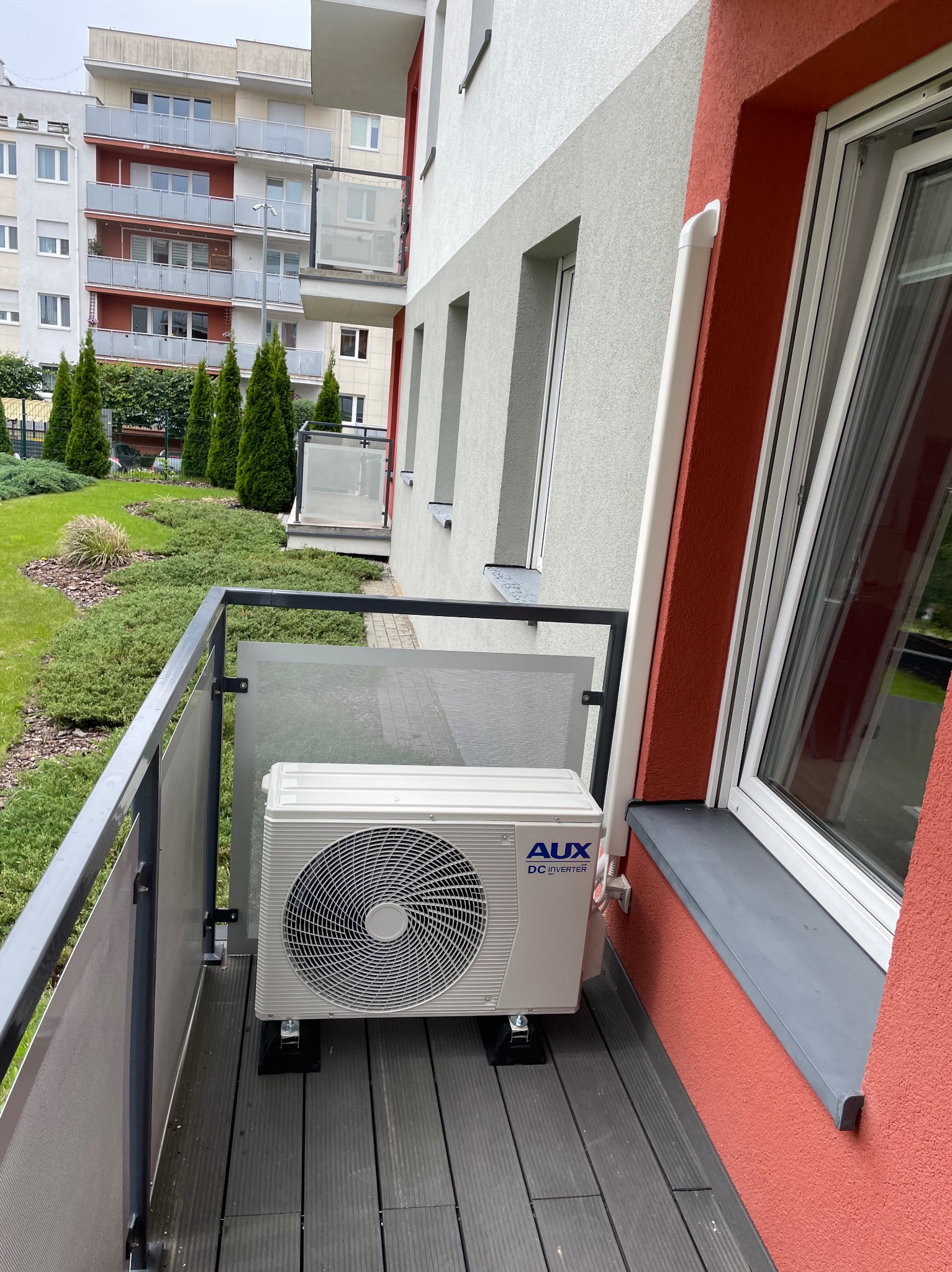 Biała jednostka zewnętrzna klimatyzacji AUX DC Inverter zamontowana na balkonie bloku mieszkalnego z widokiem na zieleń i inne budynki.