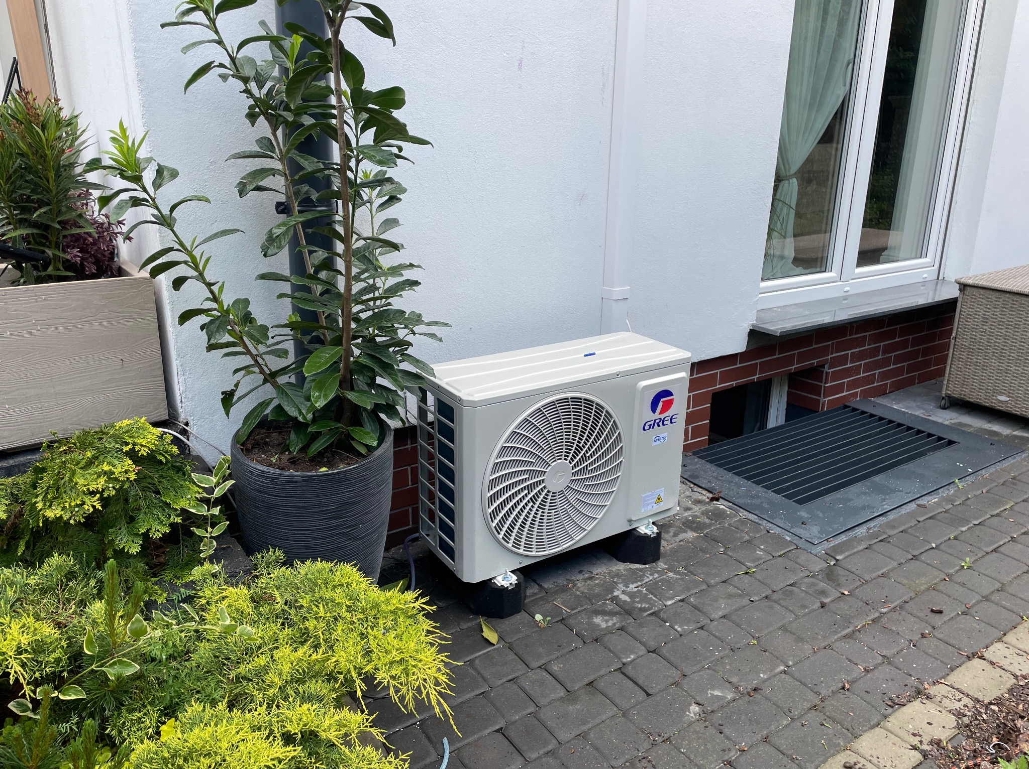 Zewnętrzna jednostka klimatyzacji Gree zamontowana przy białej ścianie domu, obok roślin w donicach i kratki okiennej piwnicy, na tle kostki brukowej.