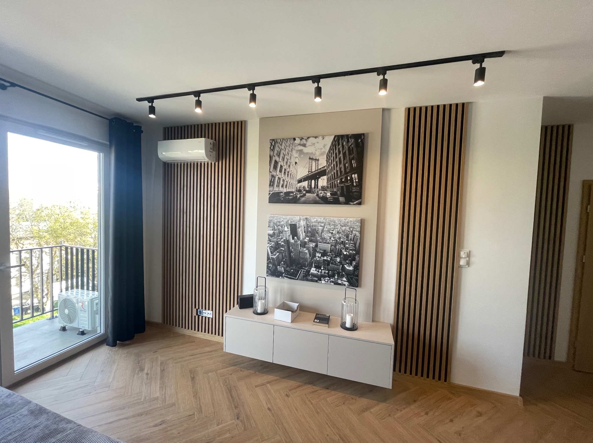 Nowoczesny salon z klimatyzacją na ścianie z drewnianymi lamelami, czarne reflektory sufitowe, obrazy Nowego Jorku i jednostka zewnętrzna klimatyzatora na balkonie.