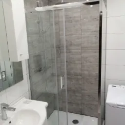 Nowoczesna łazienka z kabiną prysznicową typu walk-in, umywalką nablatową i pralką, wykończona płytkami imitującymi drewno i białymi płytkami.