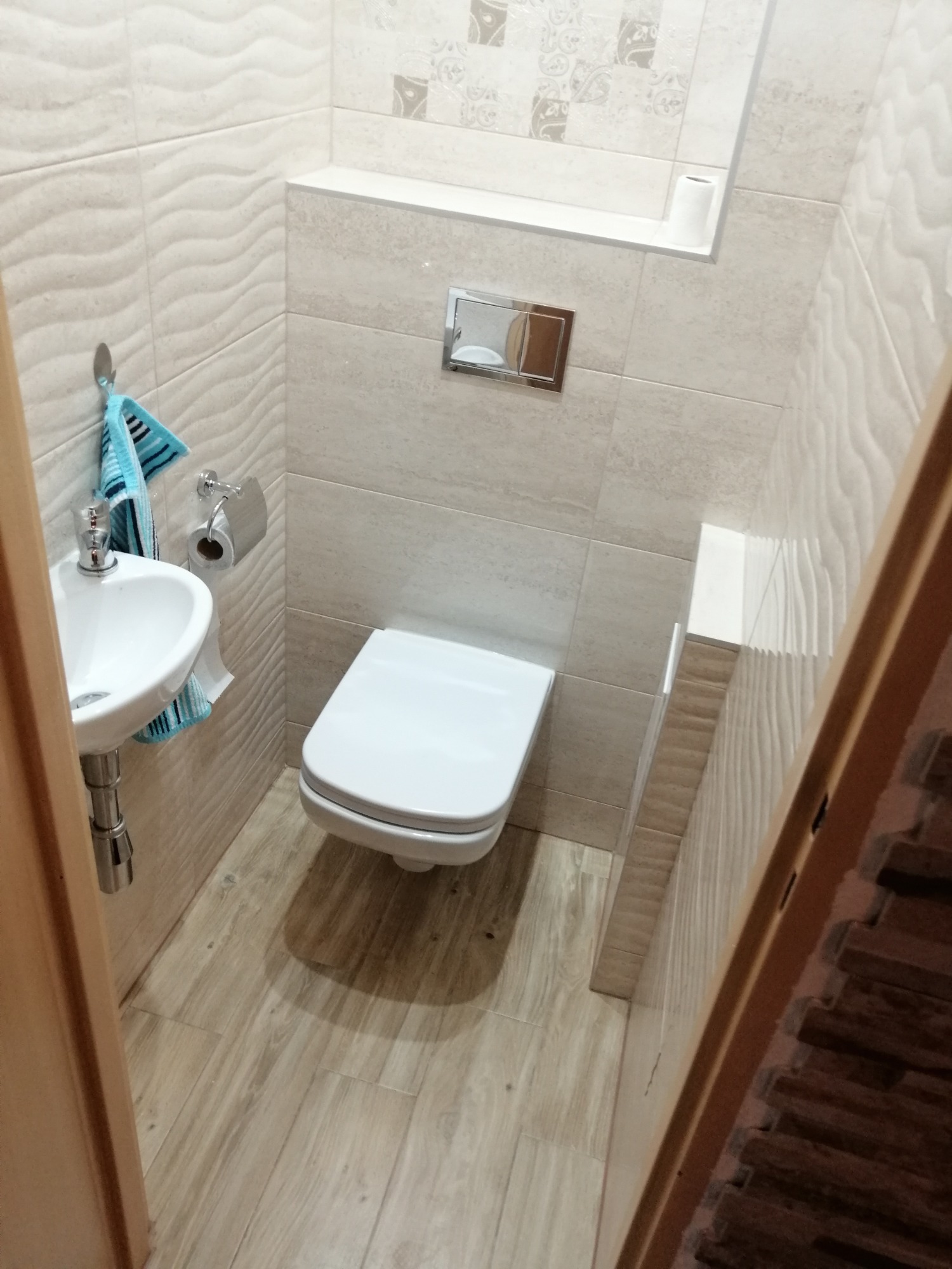 Mała, jasna łazienka z podwieszaną miską WC, umywalką narożną i beżowymi płytkami imitującymi drewno na podłodze i ścianach; widoczna niebieska ściereczka na wieszaku.