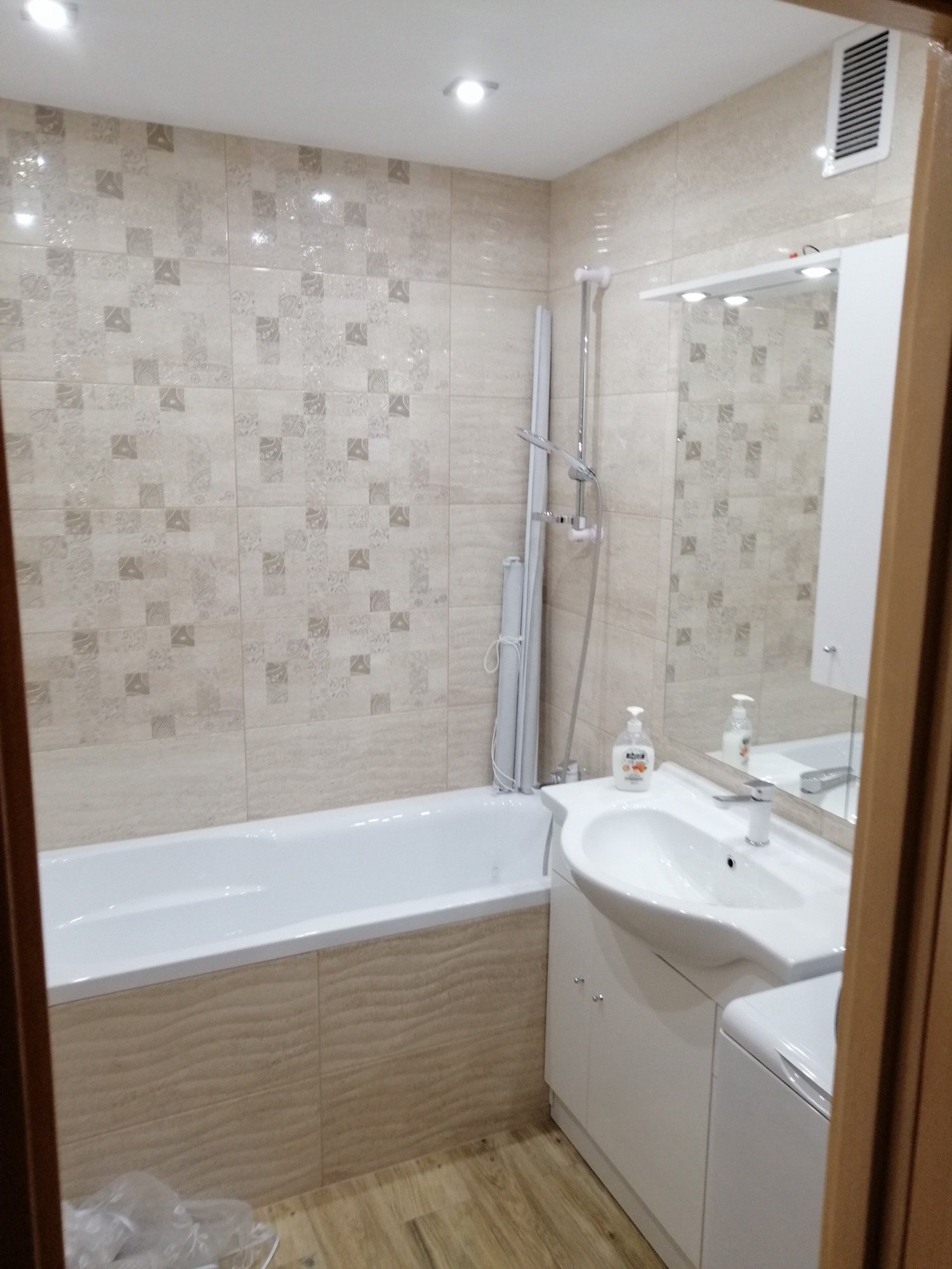 Wykończona łazienka z beżowymi płytkami, białą wanną, umywalką z szafką i prysznicem, widok zza framugi drzwi.