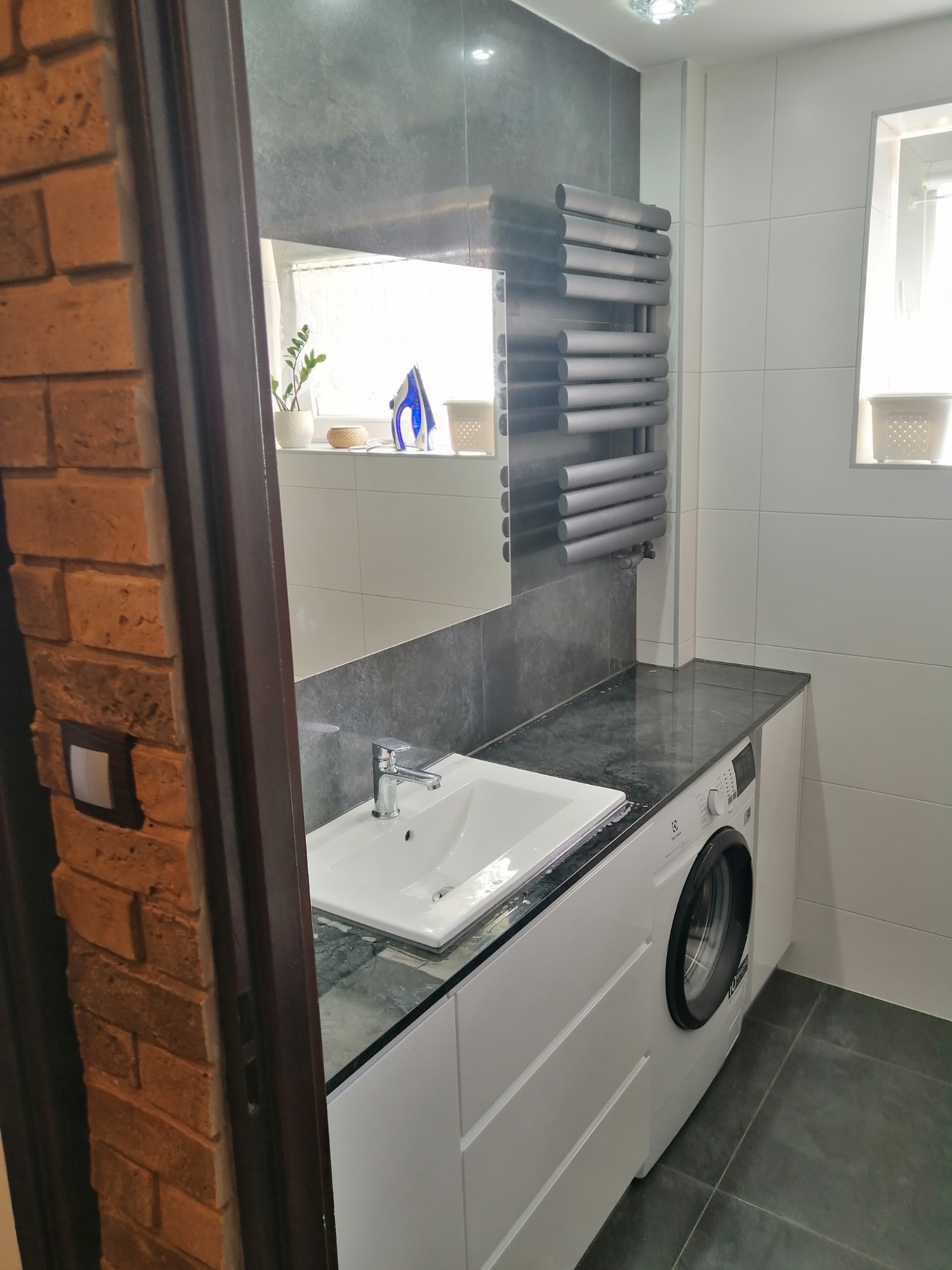 Nowoczesna łazienka z białą umywalką, pralką pod blatem z ciemnego kamienia, grzejnikiem drabinkowym i ceglaną ścianą przy drzwiach.