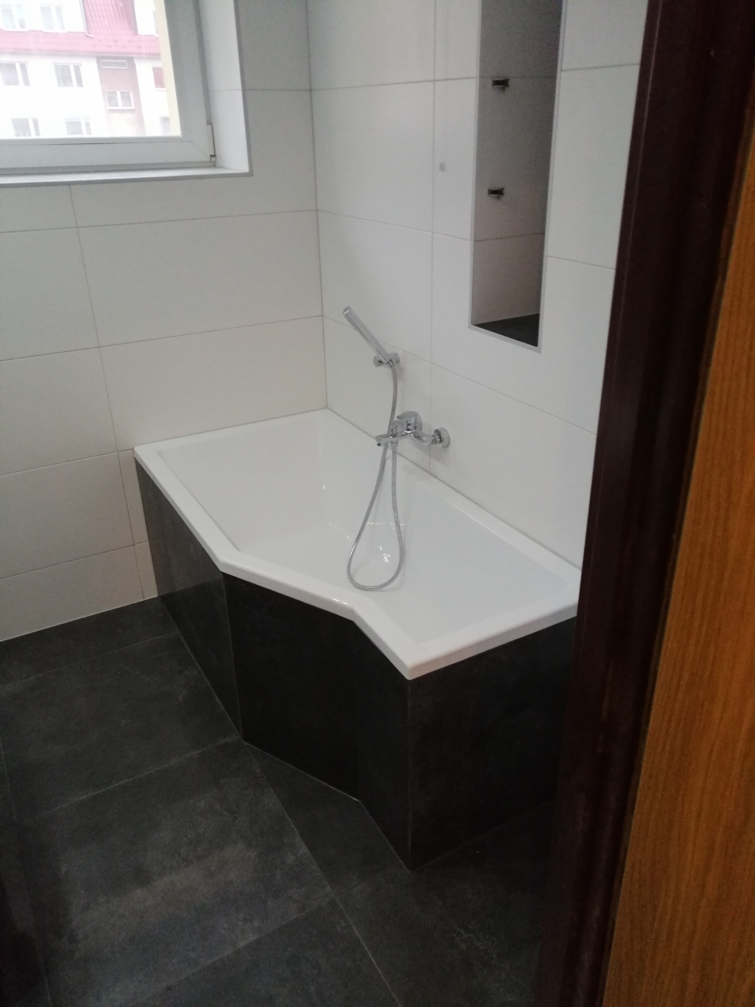 Nowoczesna łazienka z narożną wanną obudowaną ciemnymi płytkami, białe płytki na ścianach i wnęka z półkami, widok zza otwartych drzwi.