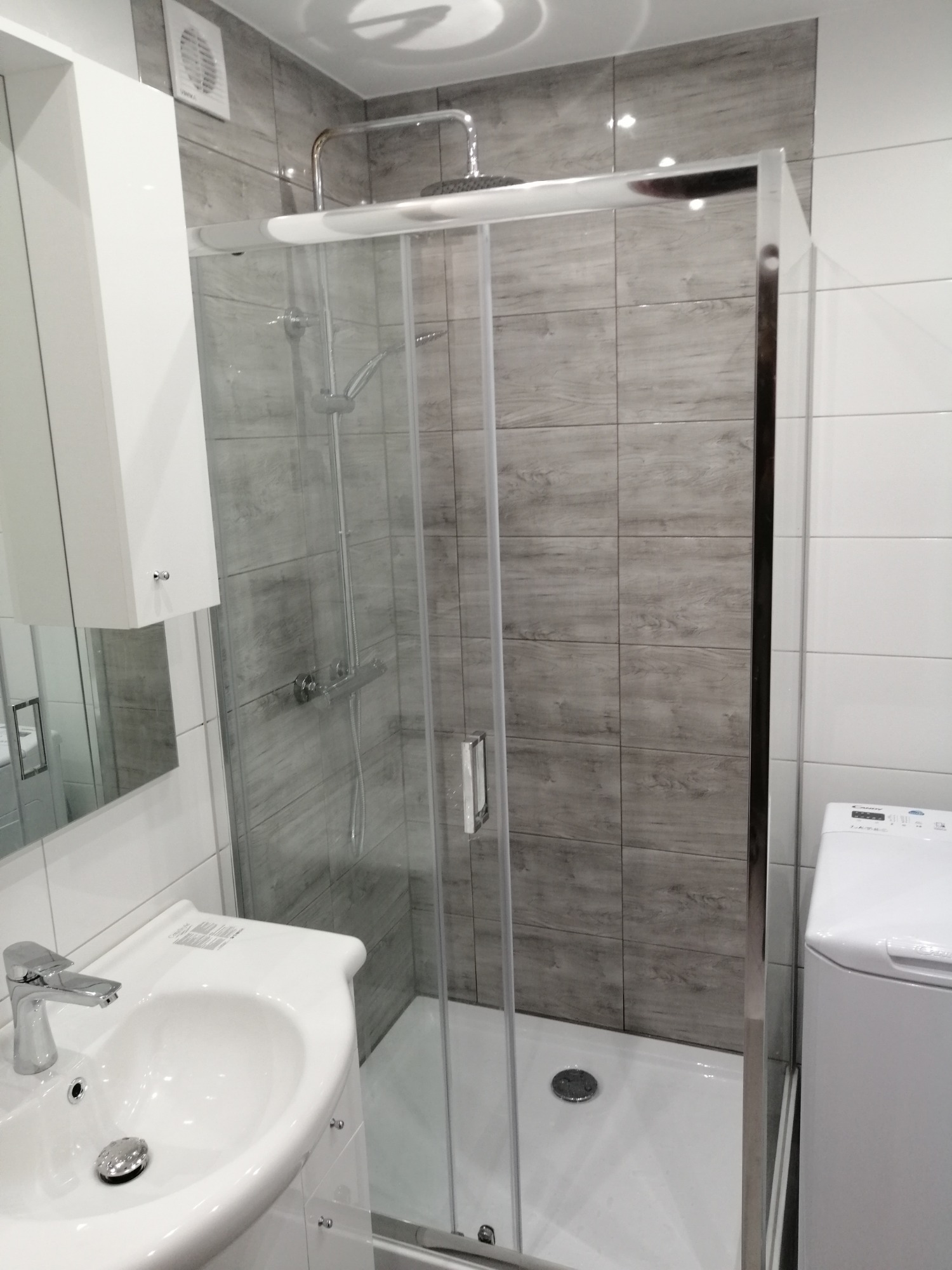 Nowoczesna łazienka z kabiną prysznicową typu walk-in, umywalką nablatową i pralką, wykończona płytkami imitującymi drewno i białymi płytkami.
